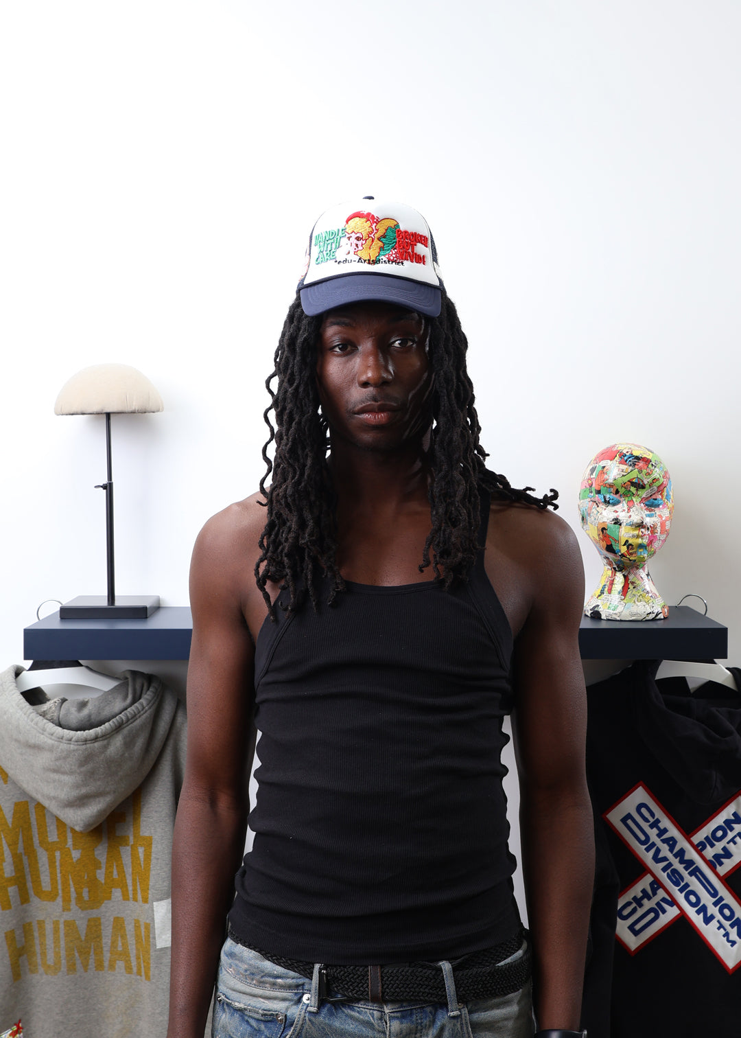 Person wearing a colorful cap and black tank top standing in front of a table with decorative items.