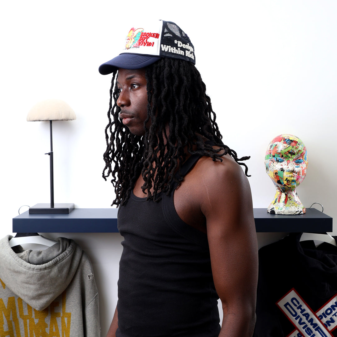 Man with dreadlocks in black tank top, cap near shelf.