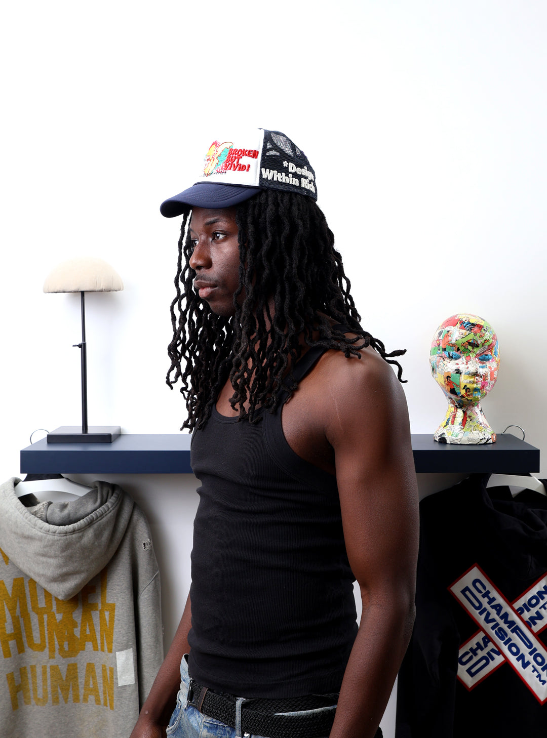Man with dreadlocks in black tank top, cap near shelf.