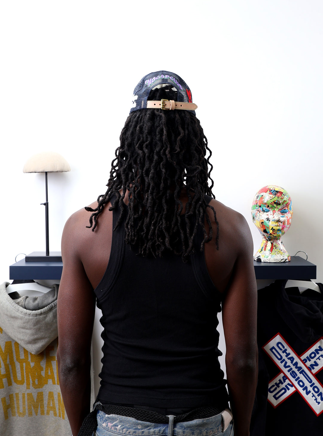 Person with dreadlocks, cap, facing art pieces on shelves.
