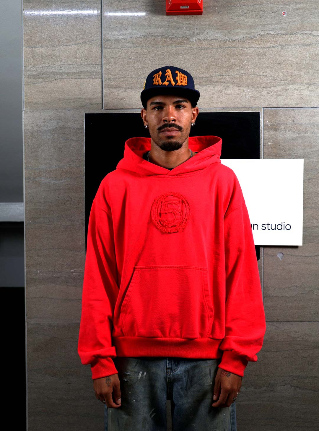 Man in red hoodie and cap standing indoors.