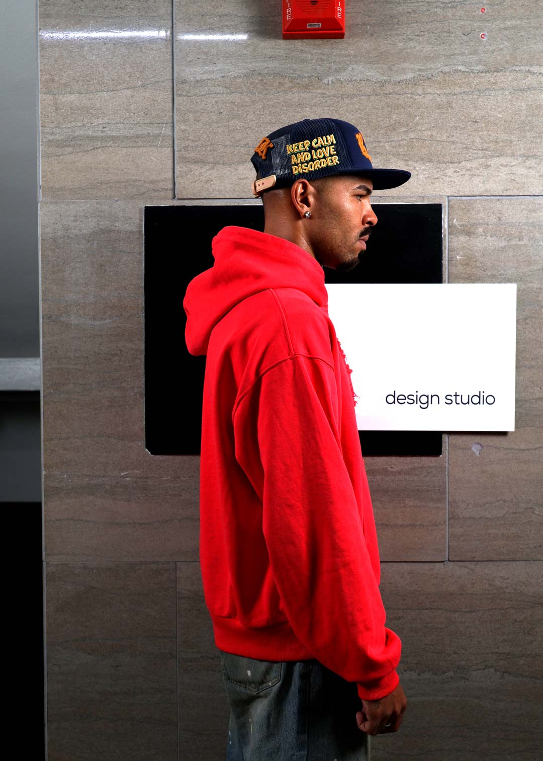 Man in red hoodie and cap near design studio sign.
