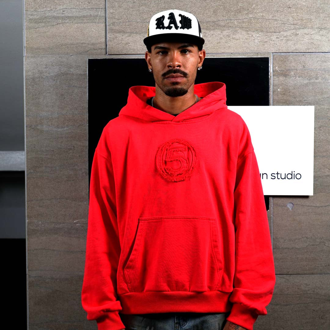 Man in red hoodie and white cap standing indoors.