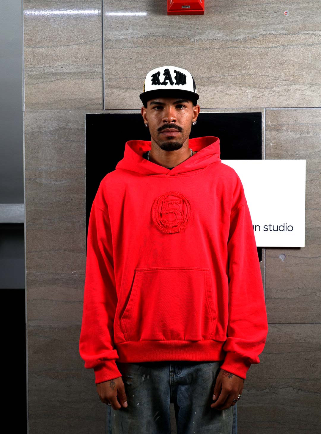 Man in red hoodie and white cap standing indoors.