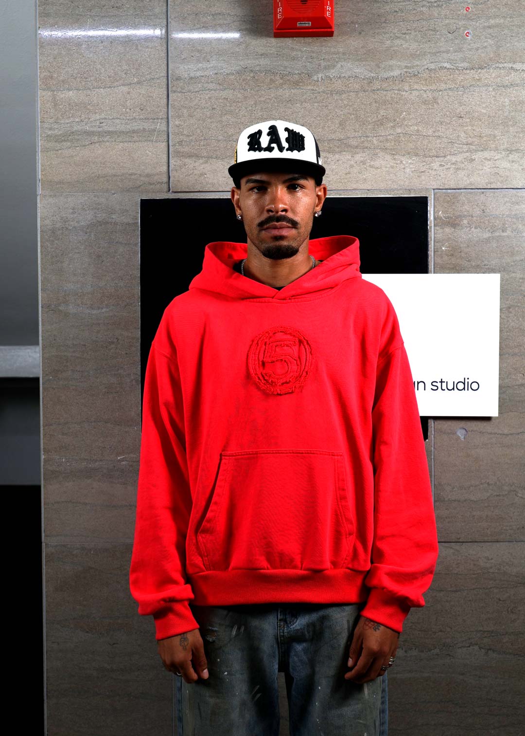 Man in red hoodie and white cap standing indoors.