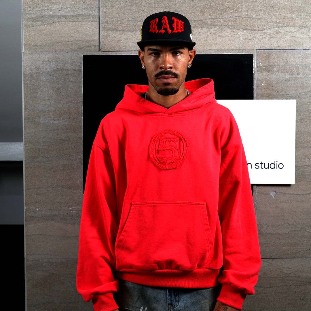 Man in red hoodie and black cap standing indoors.