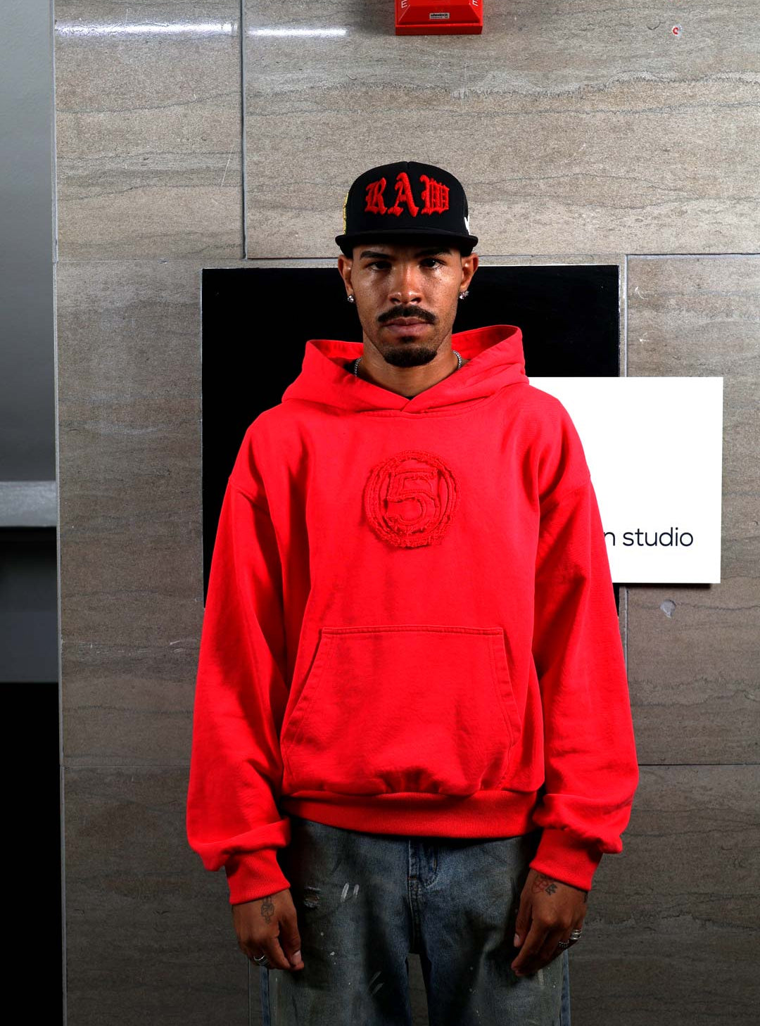 Man in red hoodie and black cap standing indoors.
