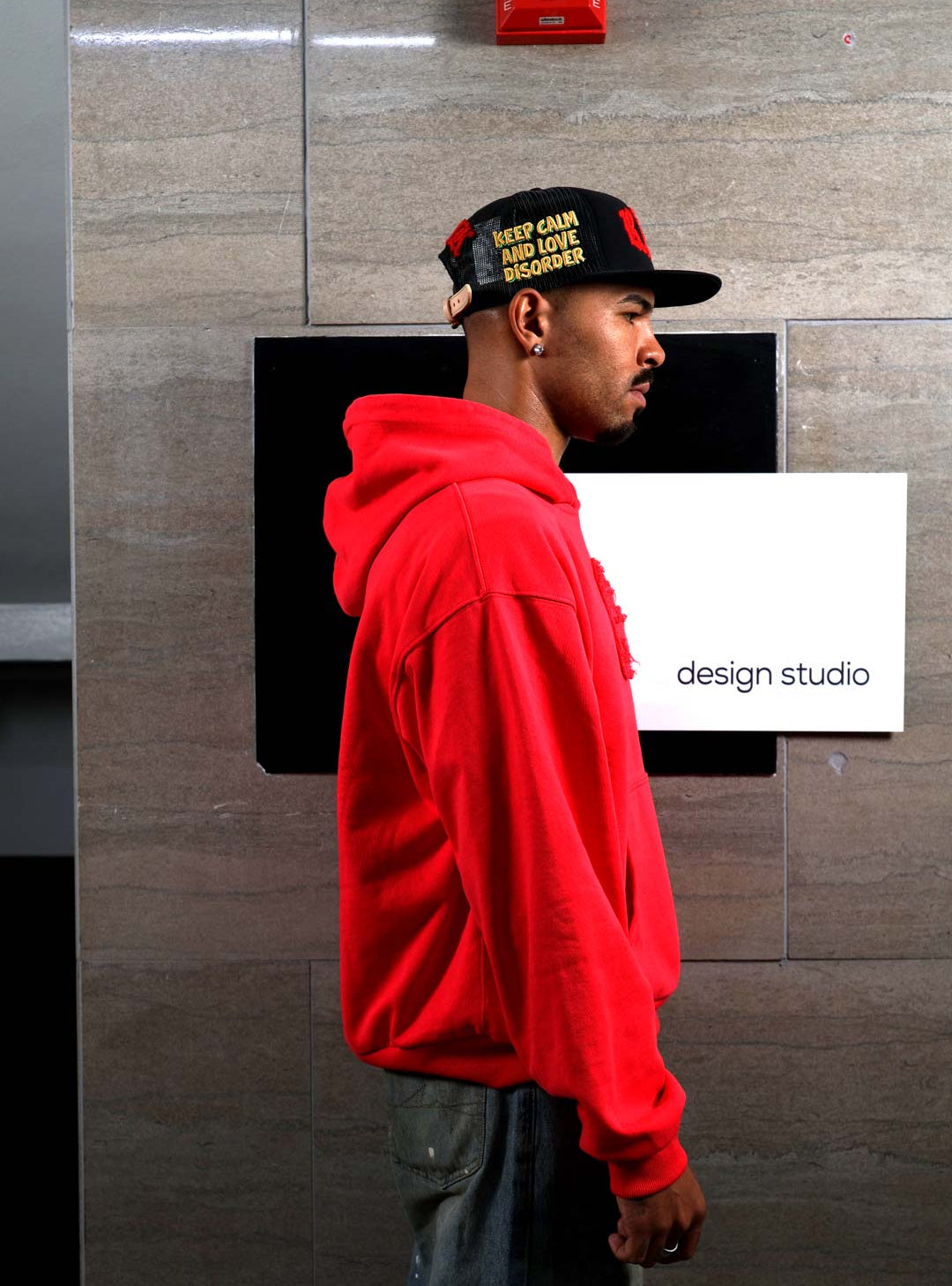Man in red hoodie and cap near design studio sign.