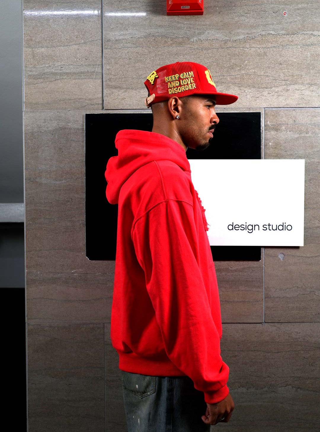 Man in red hoodie and cap near design studio sign.
