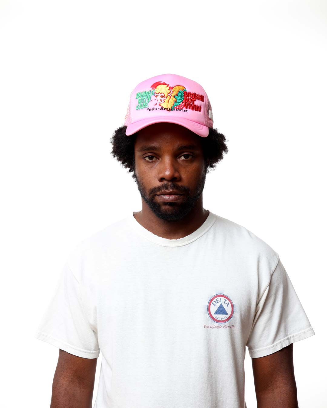 Person wearing a pink cap with colorful designs and a white t-shirt with a logo on a white background