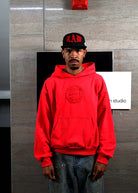 Man in red hoodie and black cap standing indoors.