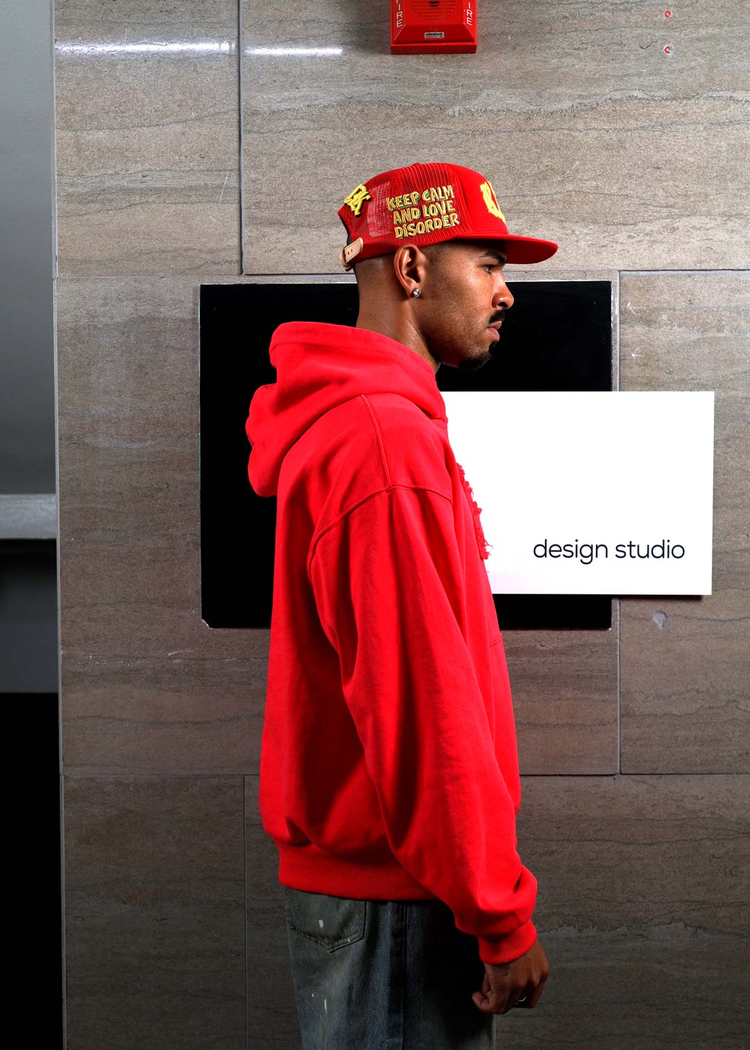 Man in red hoodie and cap near design studio sign.	